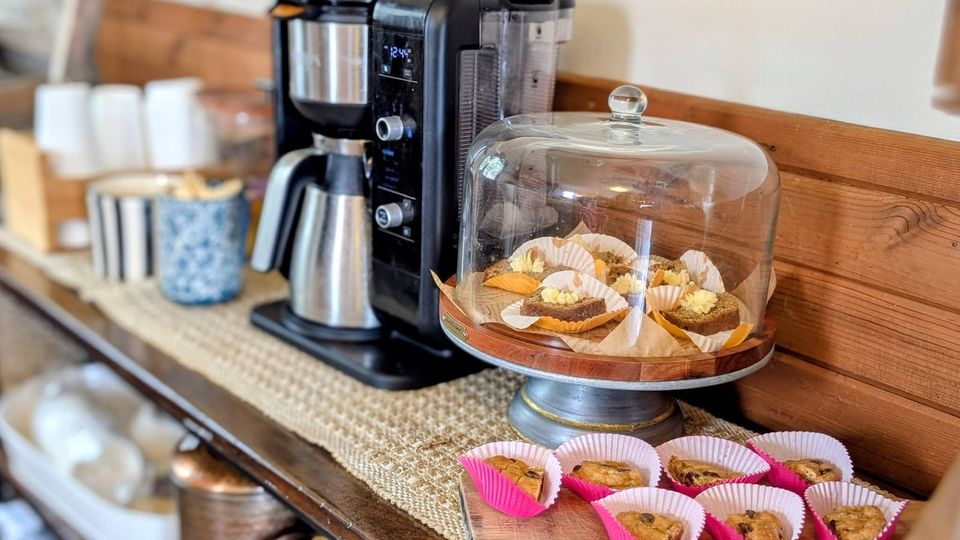 A coffee maker beside a glass cake dome of treats and muffins in pink paper cups