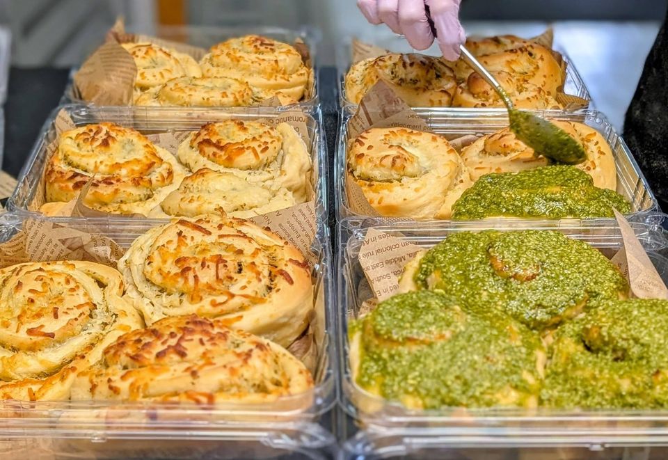 A gloved hand spooning a green herb topping onto swirled cheesy rolls in clear boxes